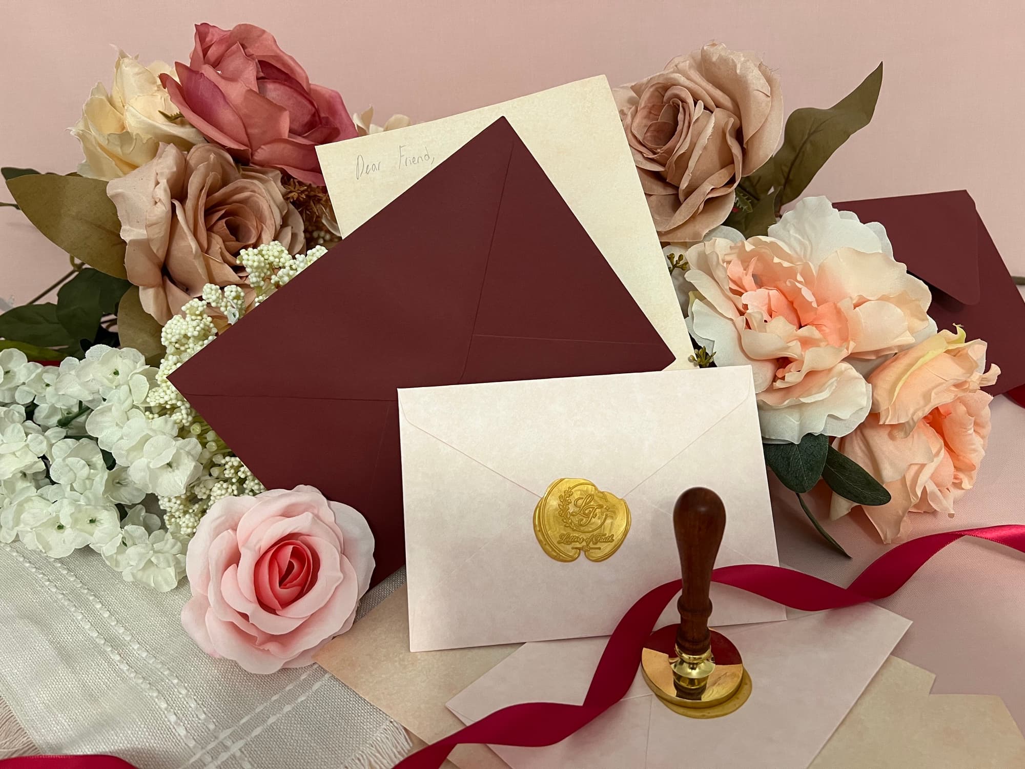 Cream and burgundy envelopes among roses, with a gold Letters of Faith wax seal, sealing stamp, and burgundy ribbon